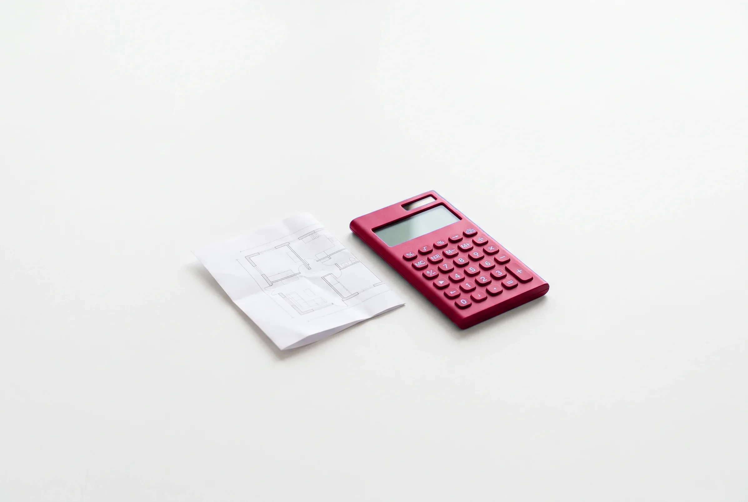 Calculadora roja junto a un plano arquitectónico doblado sobre una superficie blanca.