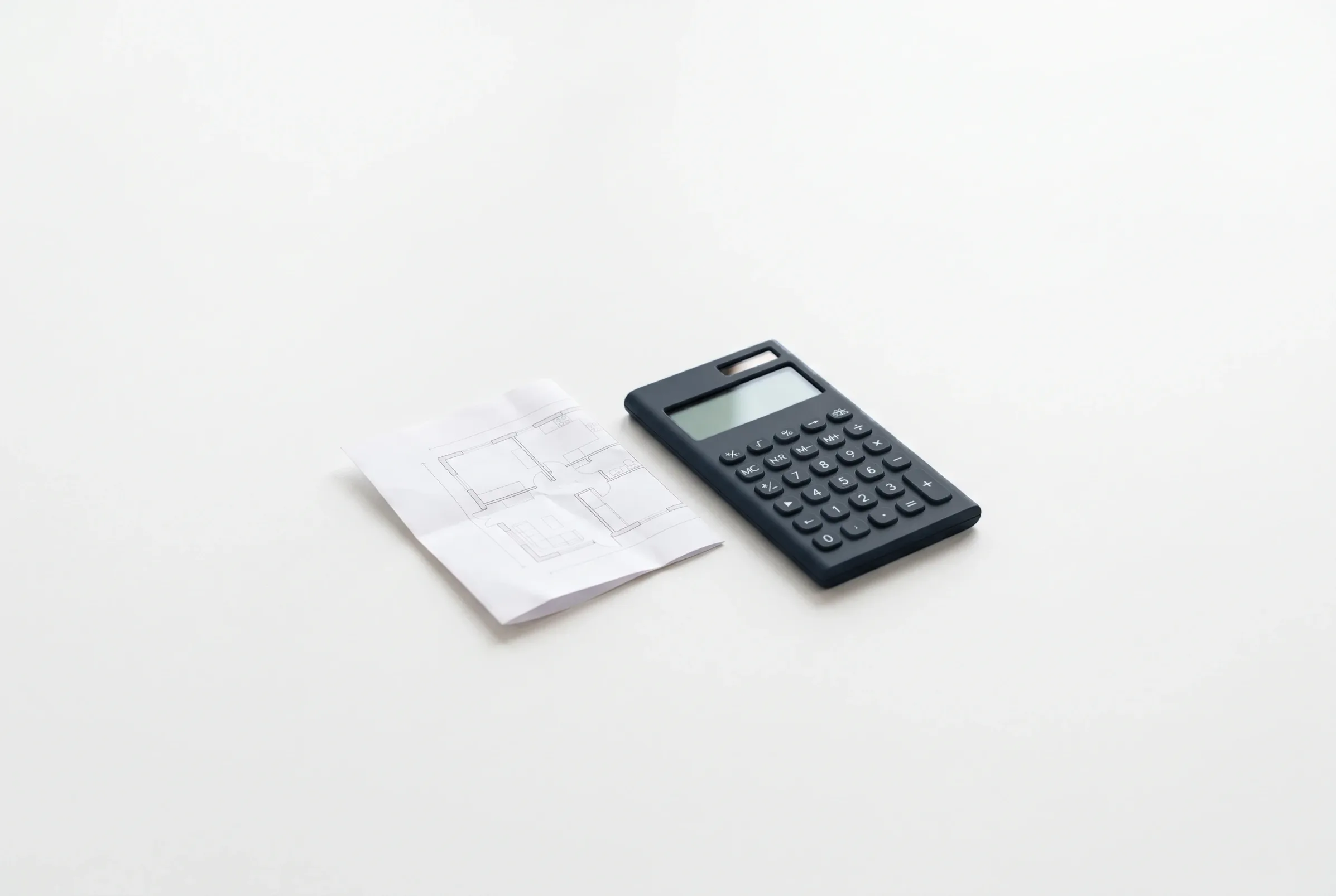 Calculadora gris pizarra junto a un plano arquitectónico doblado sobre una superficie blanca.